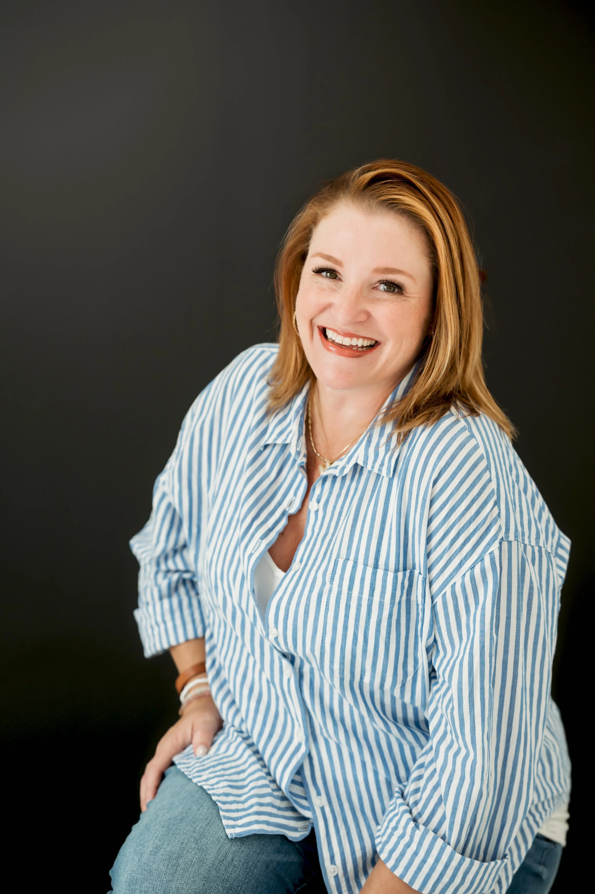 A woman with shoulder-length reddish hair smiling at the camera, wearing a blue and white striped shirt and jeans, posed against a dark background.