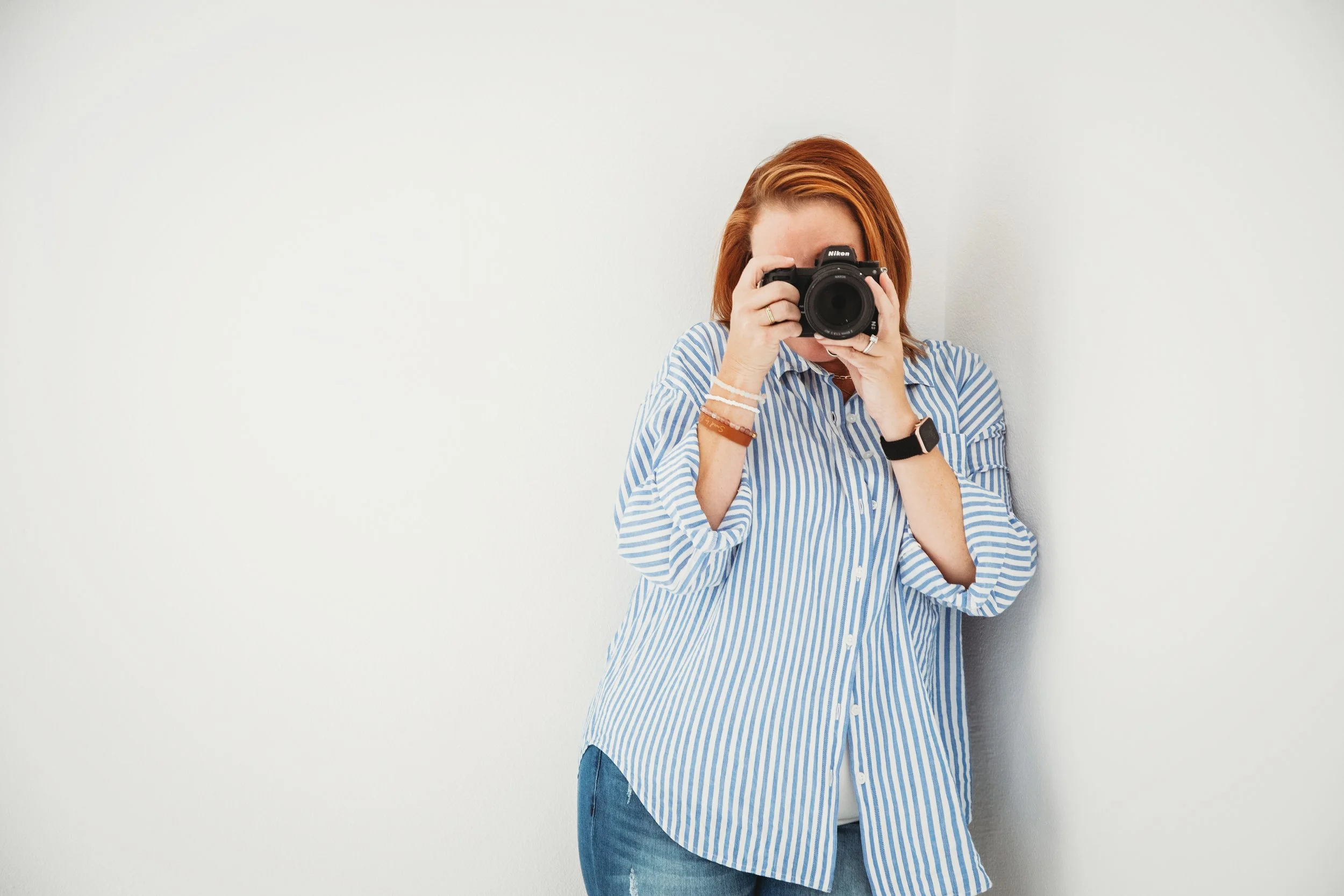 A woman with red hair wearing a blue and white striped shirt takes a photo with a camera while standing against a white wall.