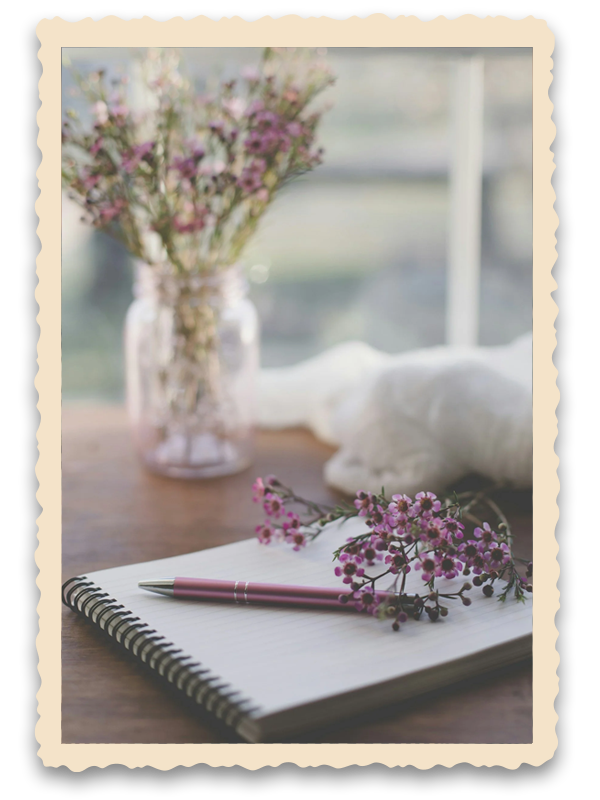 A cozy desk setup with a pink pen, a pink and purple flower, a spiral notebook, a glass vase with purple and white flowers, and a soft white blanket in the background, illuminated by natural daylight.