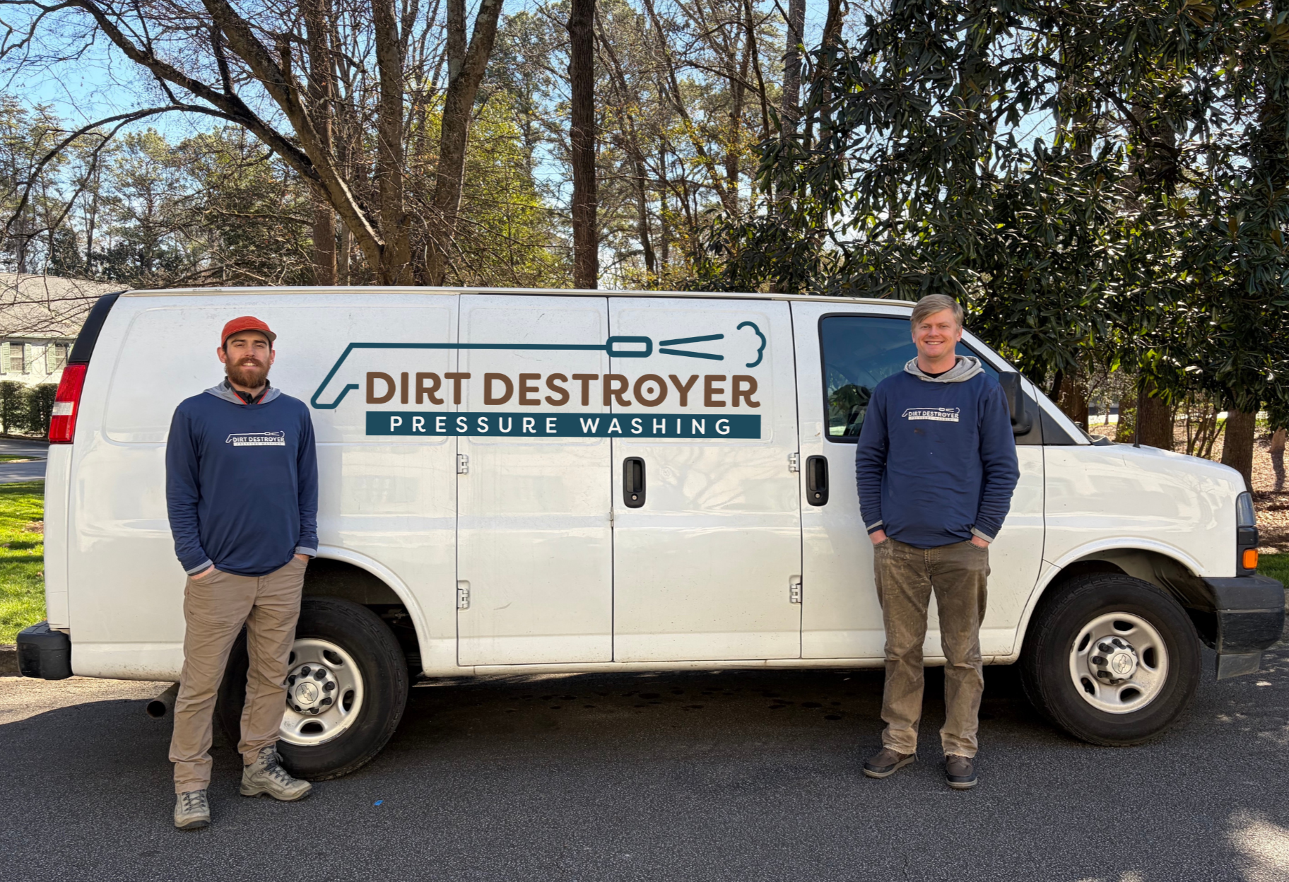 Two men standing in front of a white van with 'Dirt Destroyer Pressure Washing' logo, trees in the background, and the men wearing navy blue jackets with the same logo.