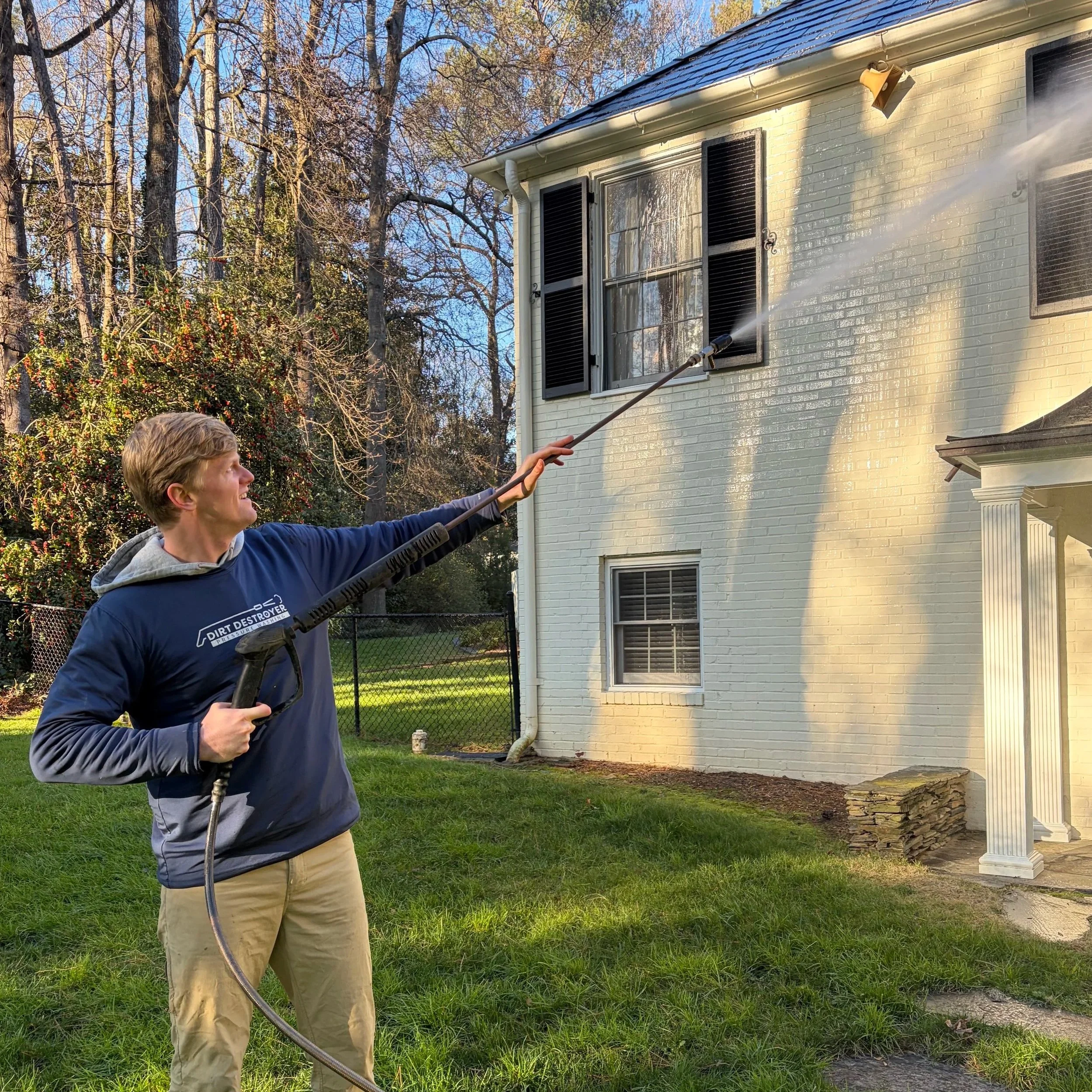 A man using a pressure washer to clean the exterior wall of a two-story house with black shutters and a white brick facade.