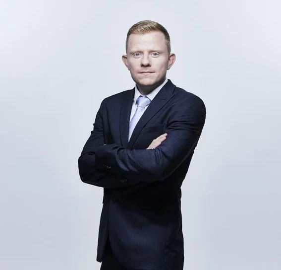 A man in a dark suit with a light blue shirt and tie, arms crossed, standing against a plain light-colored background.