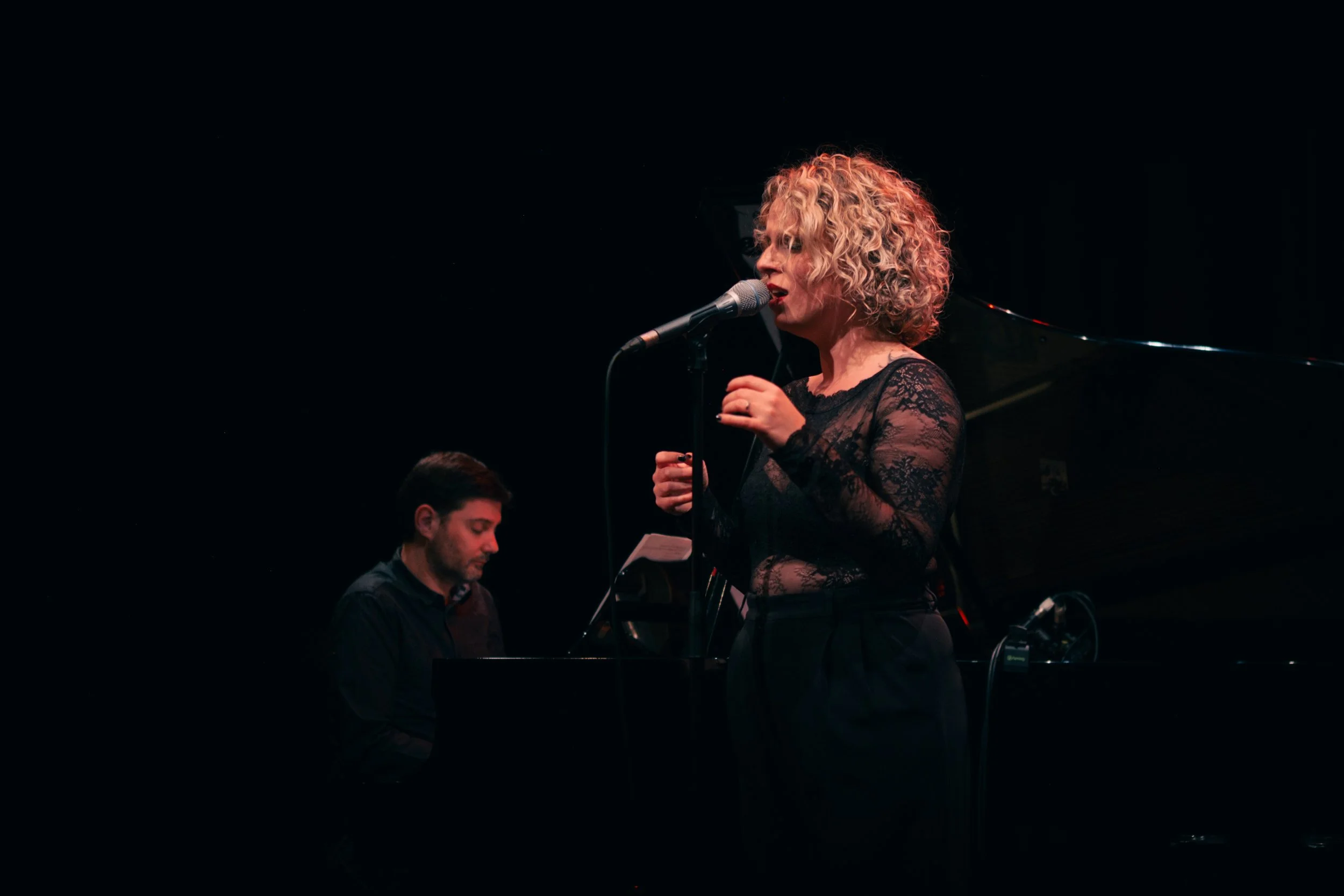 Une femme blonde à cheveux bouclés chante dans un microphone, avec un homme jouant du piano en arrière-plan, sur scène sombre.