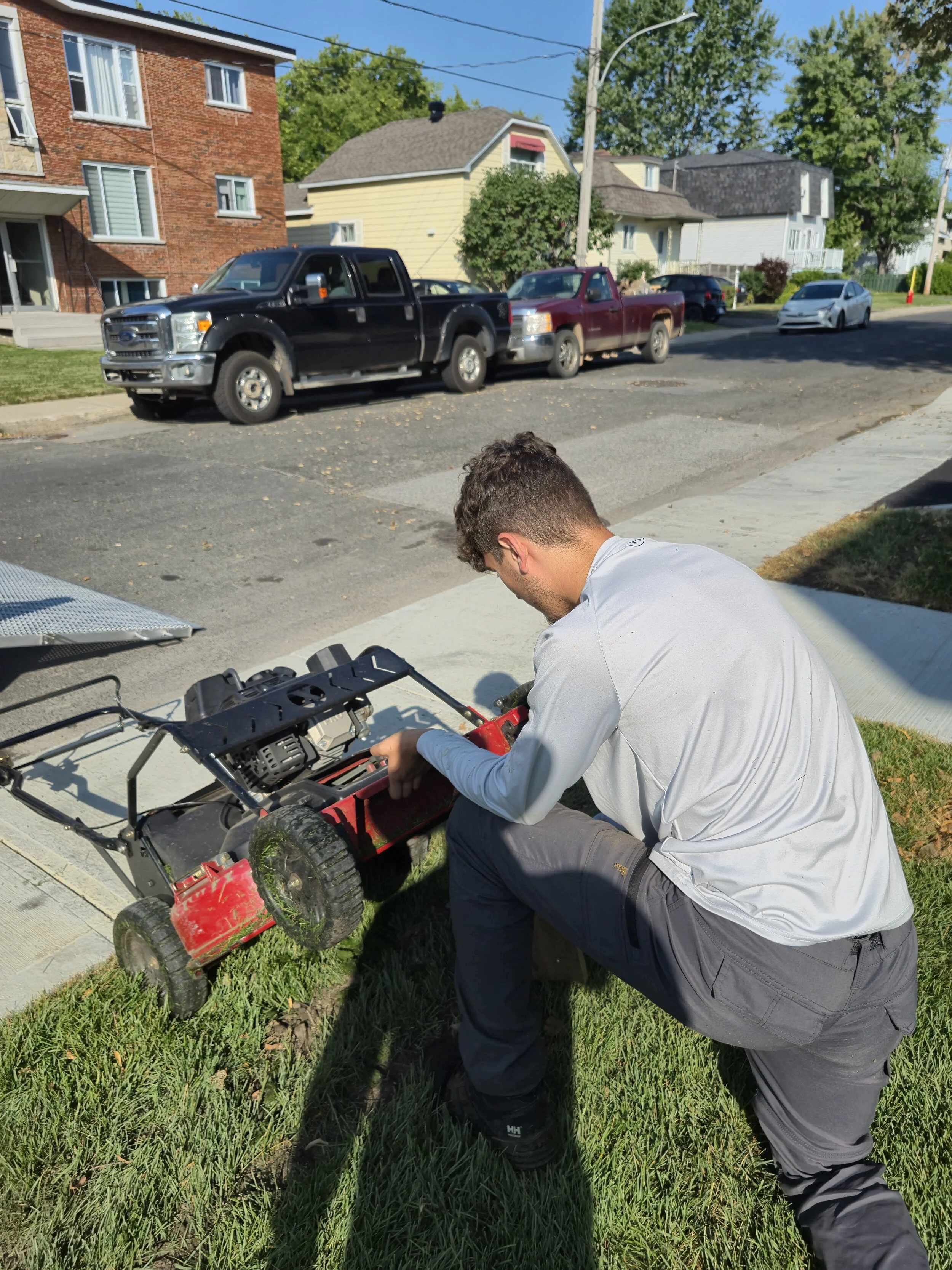 Un homme en tenue décontractée répare une tondeuse à gazon sur l'herbe près du trottoir devant des maisons résidentielles, par une journée ensoleillée.