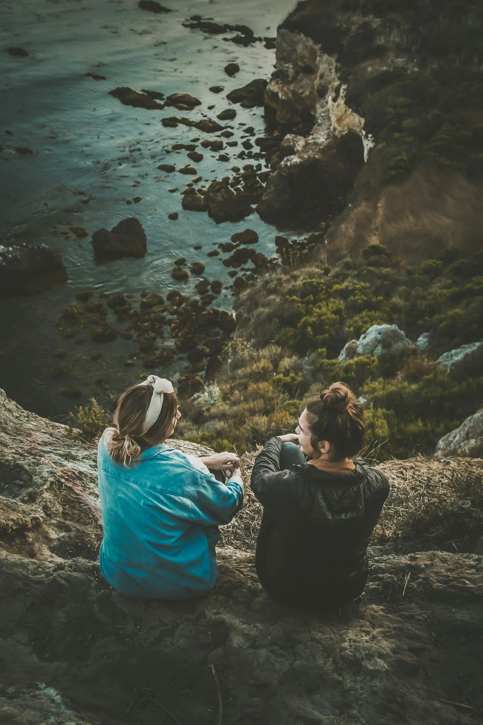 Two women sitting on a rocky cliff overlooking the ocean and rocky shoreline, engaging in conversation.
