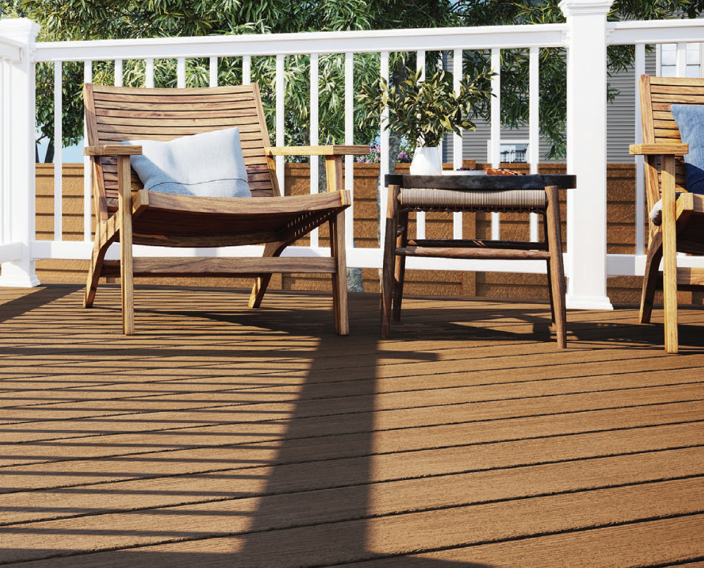 A wooden deck with two wooden chairs, one with a blue pillow, and a small table with a white vase and plant, enclosed by white railing with greenery in the background.