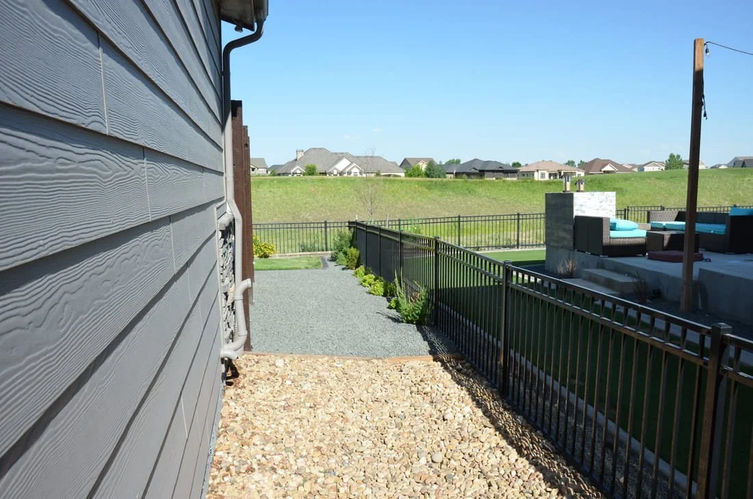 Side yard of a house with gravel and stone pathways, metal fence, and a backyard patio with outdoor furniture under a canopy.