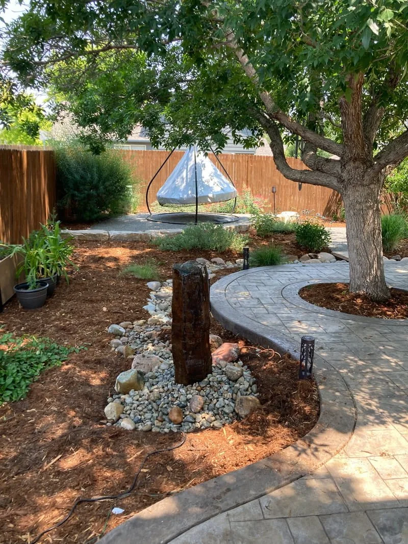 A backyard with a large green tree, a circular stone-paved pathway, a decorative stone and wood feature in mulched garden bed, and a round fabric canopy swing on a concrete platform in the background.