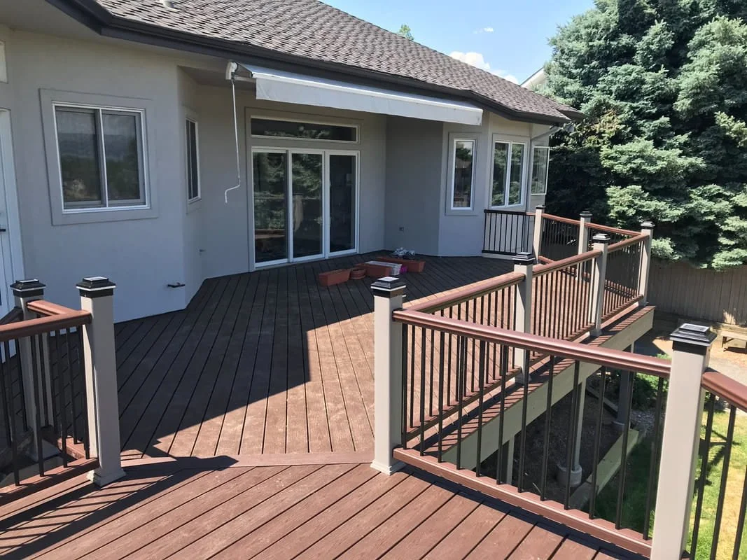 Backyard wooden deck attached to a house with sliding glass doors and windows, surrounded by a railing, with trees in the background.
