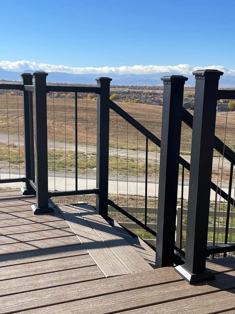 A black metal railing on a wooden deck overlooking a rural landscape with fields, a few houses, and mountains in the distance under a clear blue sky.