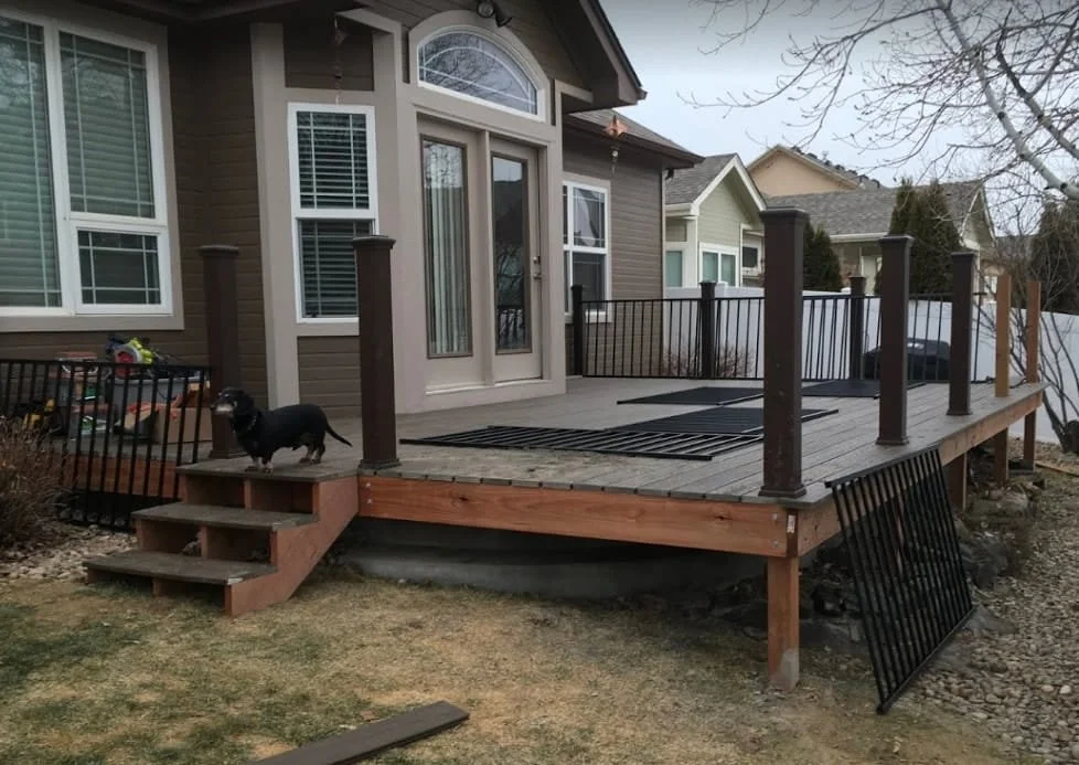 A wooden backyard deck with black railings, attached to a house with gray siding and white-framed windows. A black dog with a collar is standing on the stairs leading up to the deck, and there's a small section of a gate leaning against the deck's su