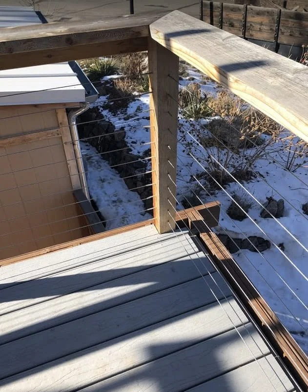 Close-up of a wooden deck with a safety railing, overlooking a snowy backyard with rocks and plants, seen from an elevated position.
