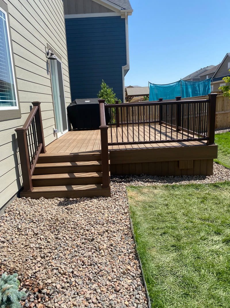 Backyard deck with wooden stairs, black railing, a grill, and a view of neighboring houses with a blue safety net in the yard.