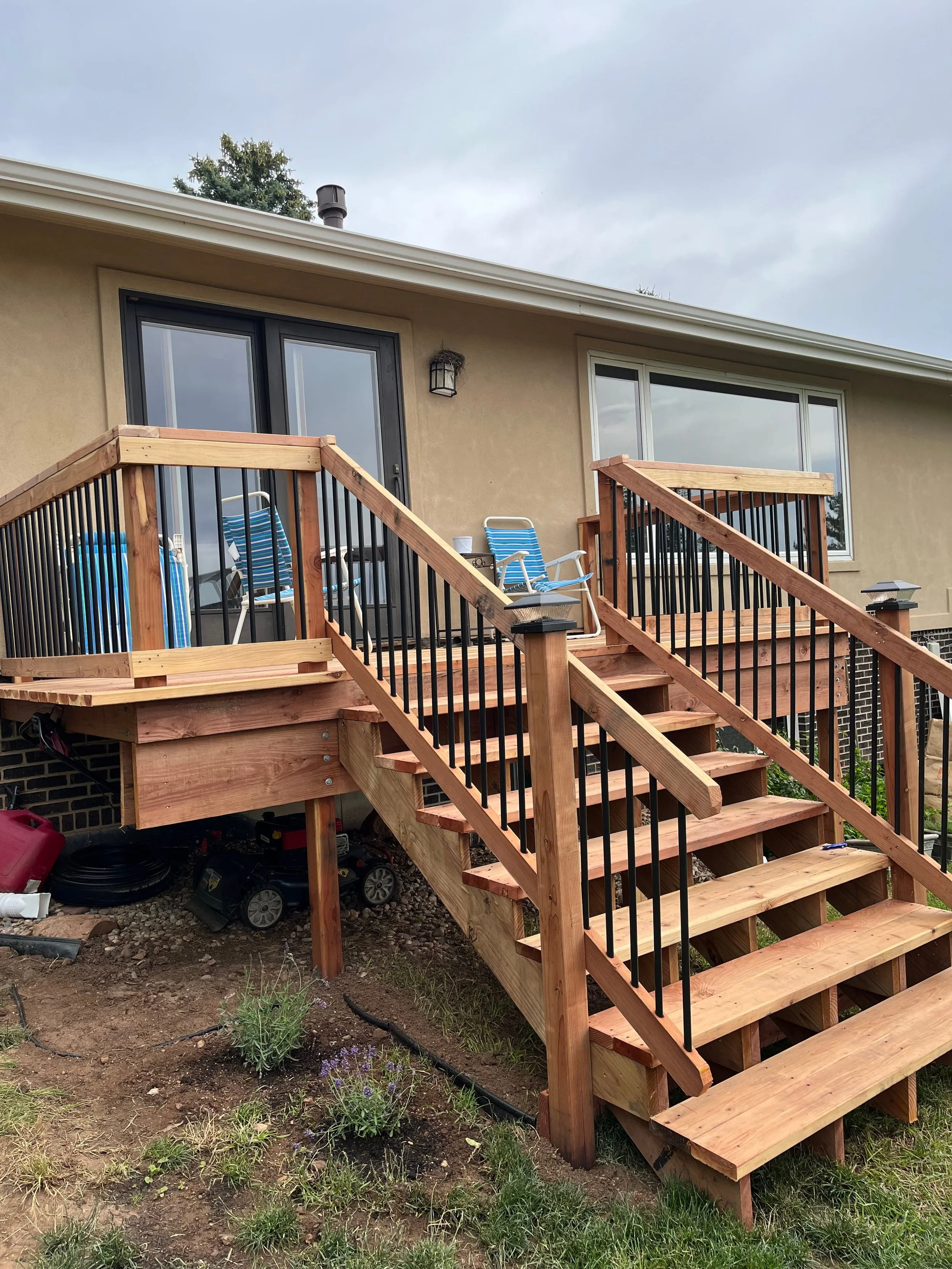 Backyard deck under construction with new wooden stairs and black metal railings, attached to a beige house with sliding glass door and window, with a cloudy sky in the background.