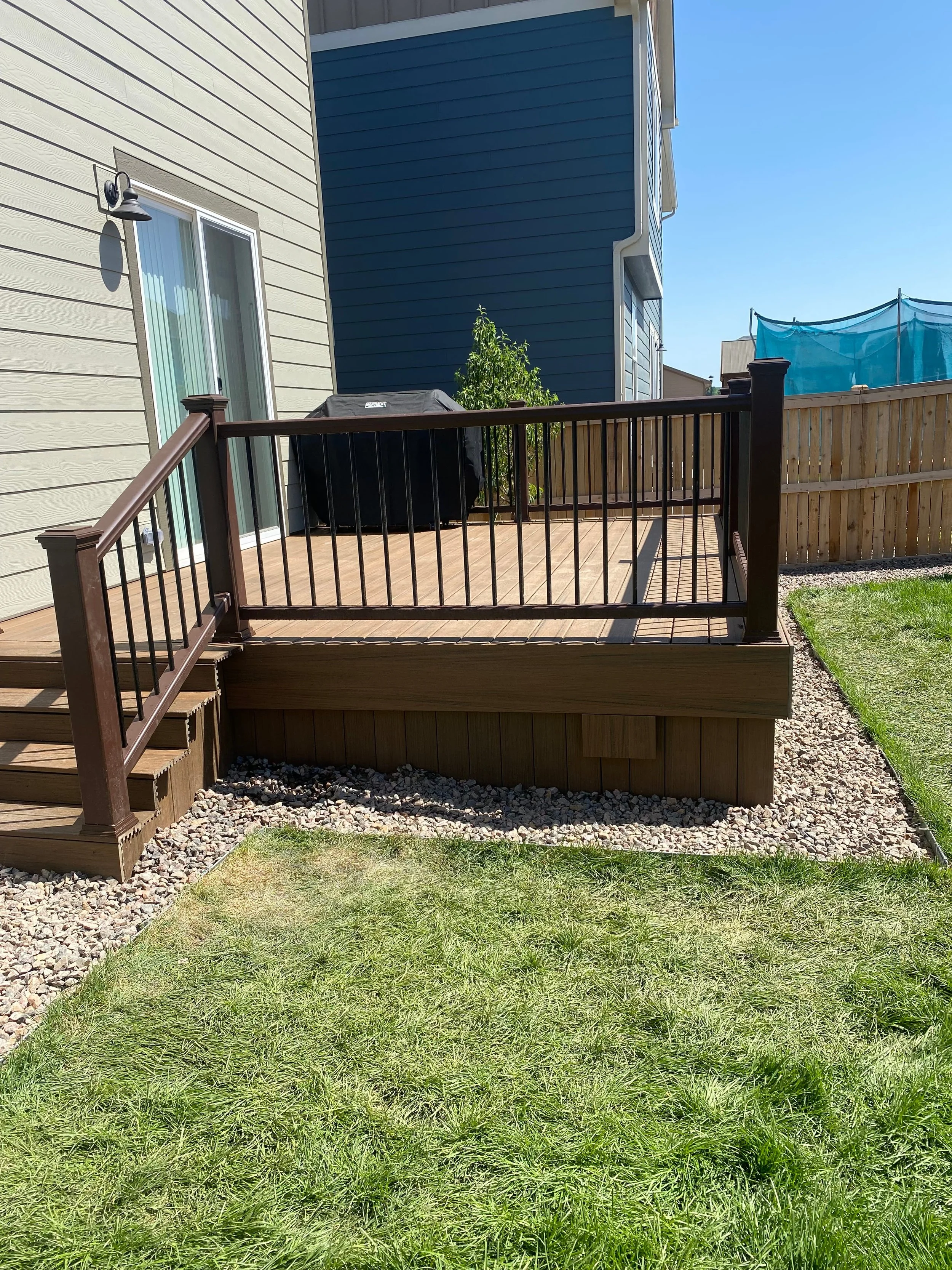 Backyard deck with wooden railing, steps, and grassy yard, adjacent to a house with beige and blue siding, and a fenced-in area with a trampoline in the background.