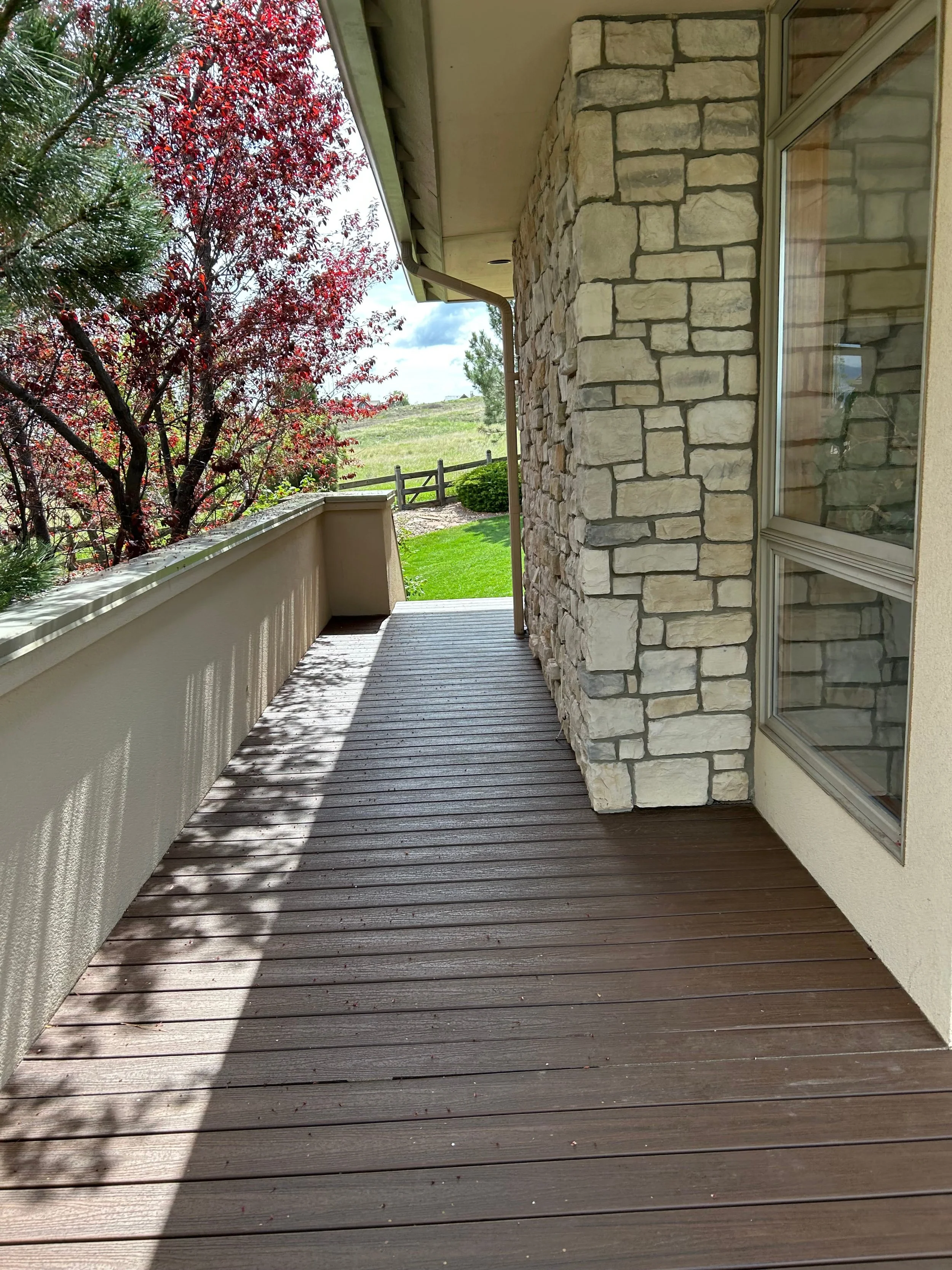 View of a wooden porch with a stone wall, windows, a tree with red leaves, and a fenced yard with green grass and a cloudy sky