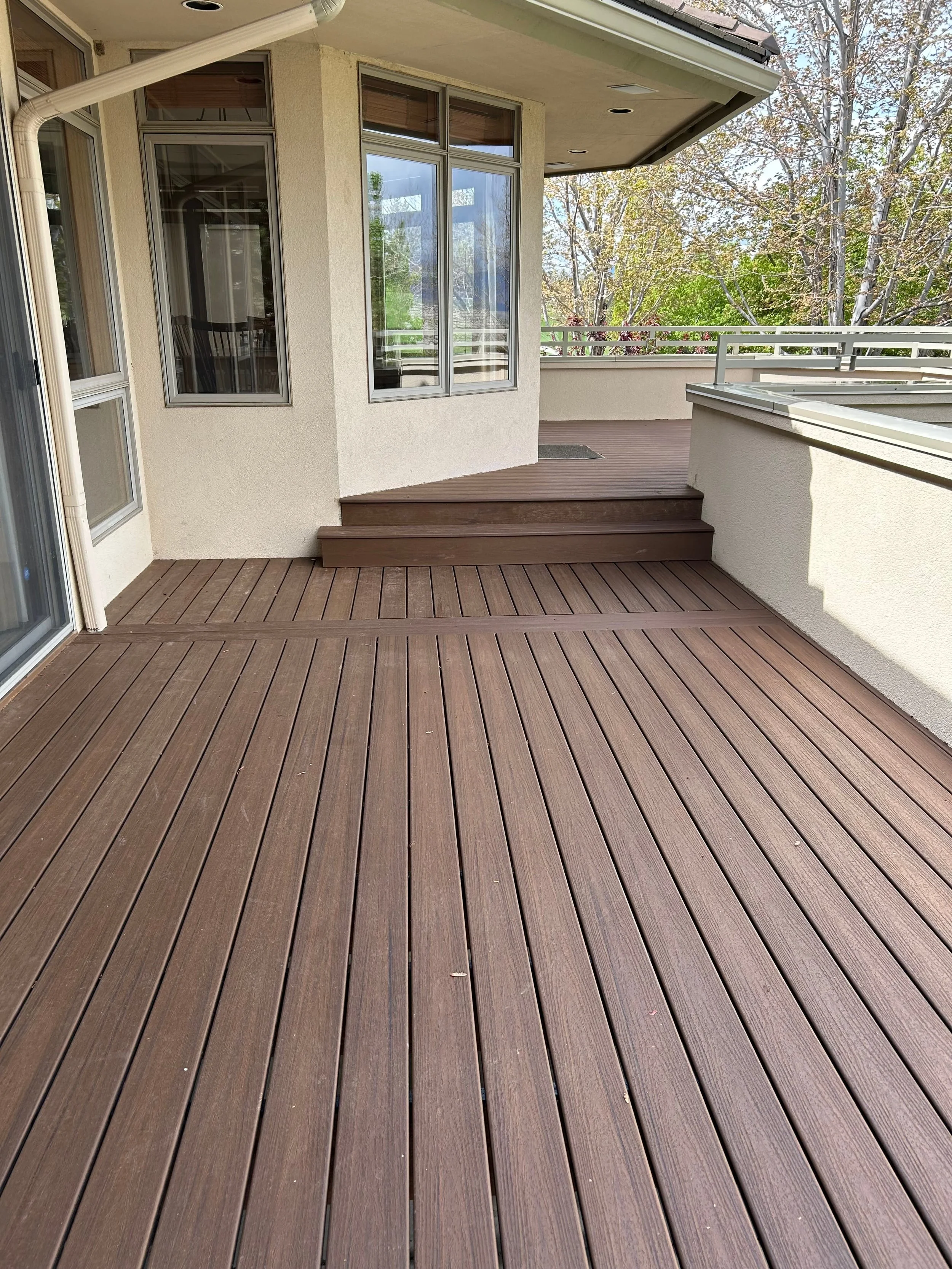 View of a spacious outdoor balcony with wooden deck flooring, staircase, glass windows, and a partial view of trees and sky in the background.