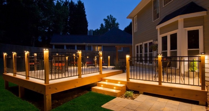 Night view of a backyard deck with lighting and stairs, attached to a house with large windows.