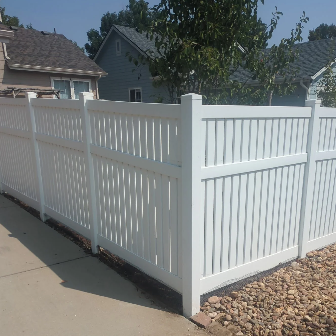 White vinyl privacy fence installed along a sidewalk with houses and trees in the background.