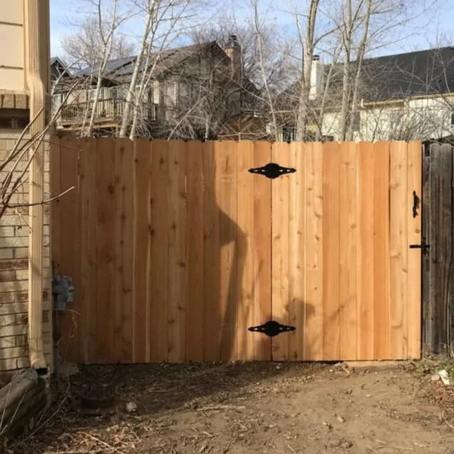 New wooden backyard gate with decorative black hinges and latch, surrounded by a dirt ground and neighboring houses and trees in background.