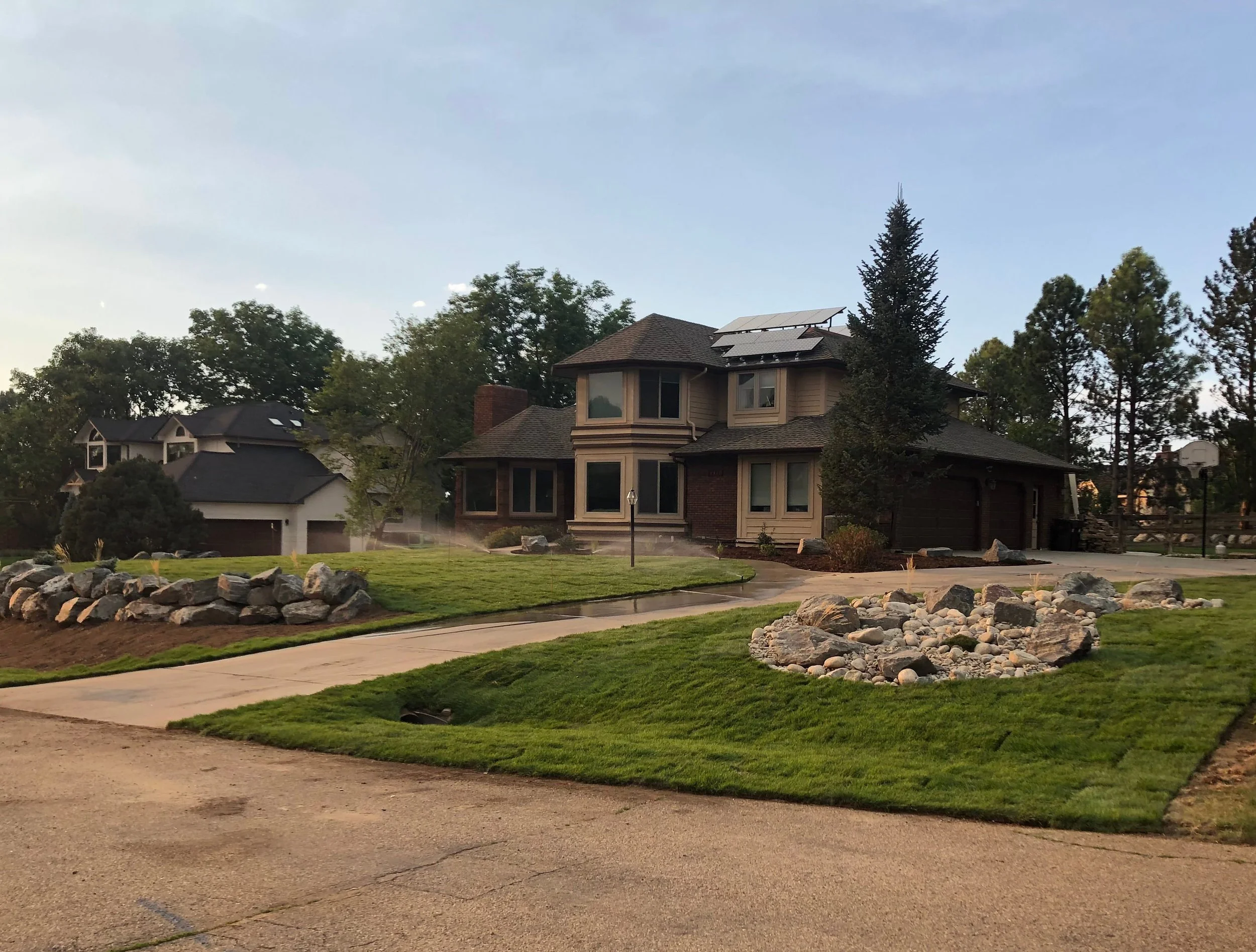 A large two-story house with solar panels on the roof, surrounded by a well-maintained lawn, trees, rocks, and rocks arranged in a landscape design. There is a sidewalk and driveway in the foreground and a basketball hoop in the driveway to the right