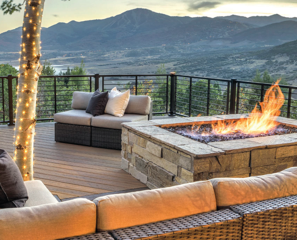 Outdoor patio with a fire pit, sectional sofa, pillows, and string lights wrapped around a tree. Colorado Mountains in the background 