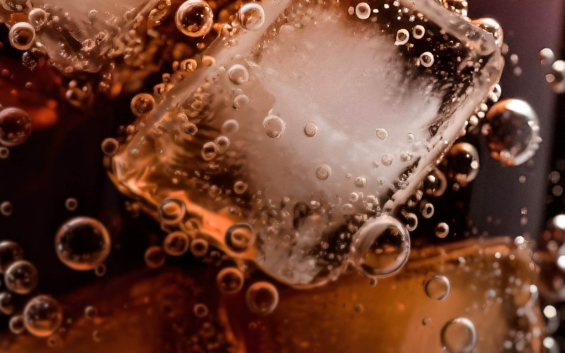 Close-up of a clear ice cube with bubbles inside, surrounded by smaller bubbles, with warm amber and dark background.