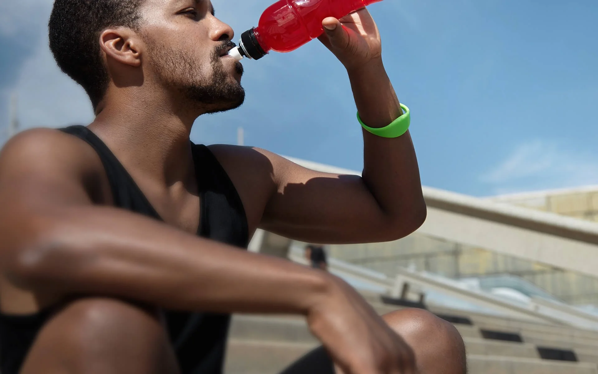A man with short hair and a beard is drinking a red sports drink from a bottle. He is wearing a black sleeveless athletic shirt and a green wristband, sitting outdoors against a bright sky and modern architecture.