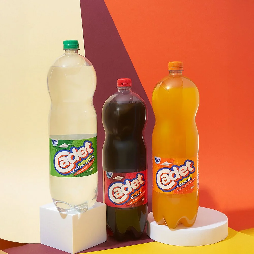 Three bottles of Adet soda in lemon & lime, cola, and orange flavors, placed on white blocks against a colorful, geometric background.