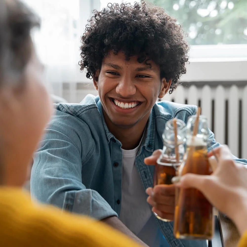 Young man with curly hair smiling and holding a bottle of beer, clinking glasses with someone in a social setting.