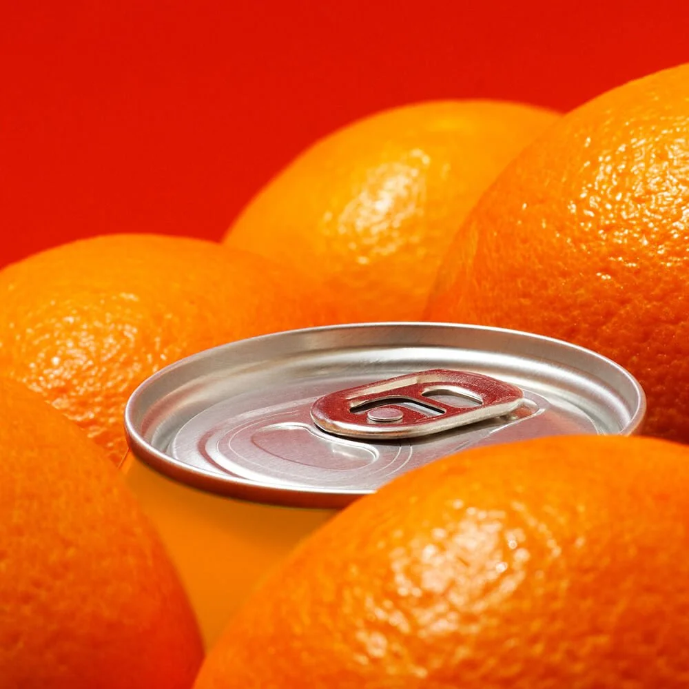A close-up of a soda can surrounded by multiple oranges against a red background.