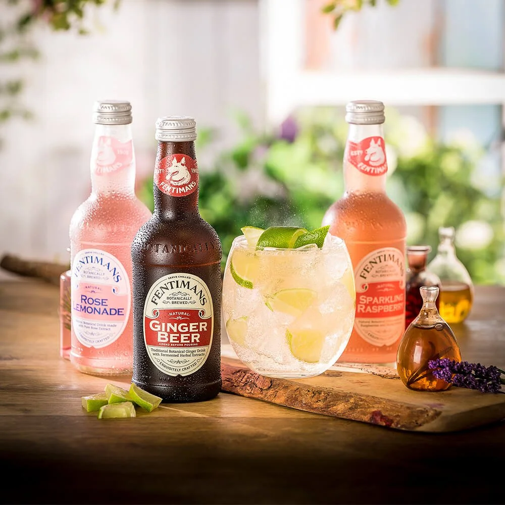 Three bottles of Fentimans flavored beverages, a glass of lemonade with lime wedges, and small bottles with lavender on a wooden surface.