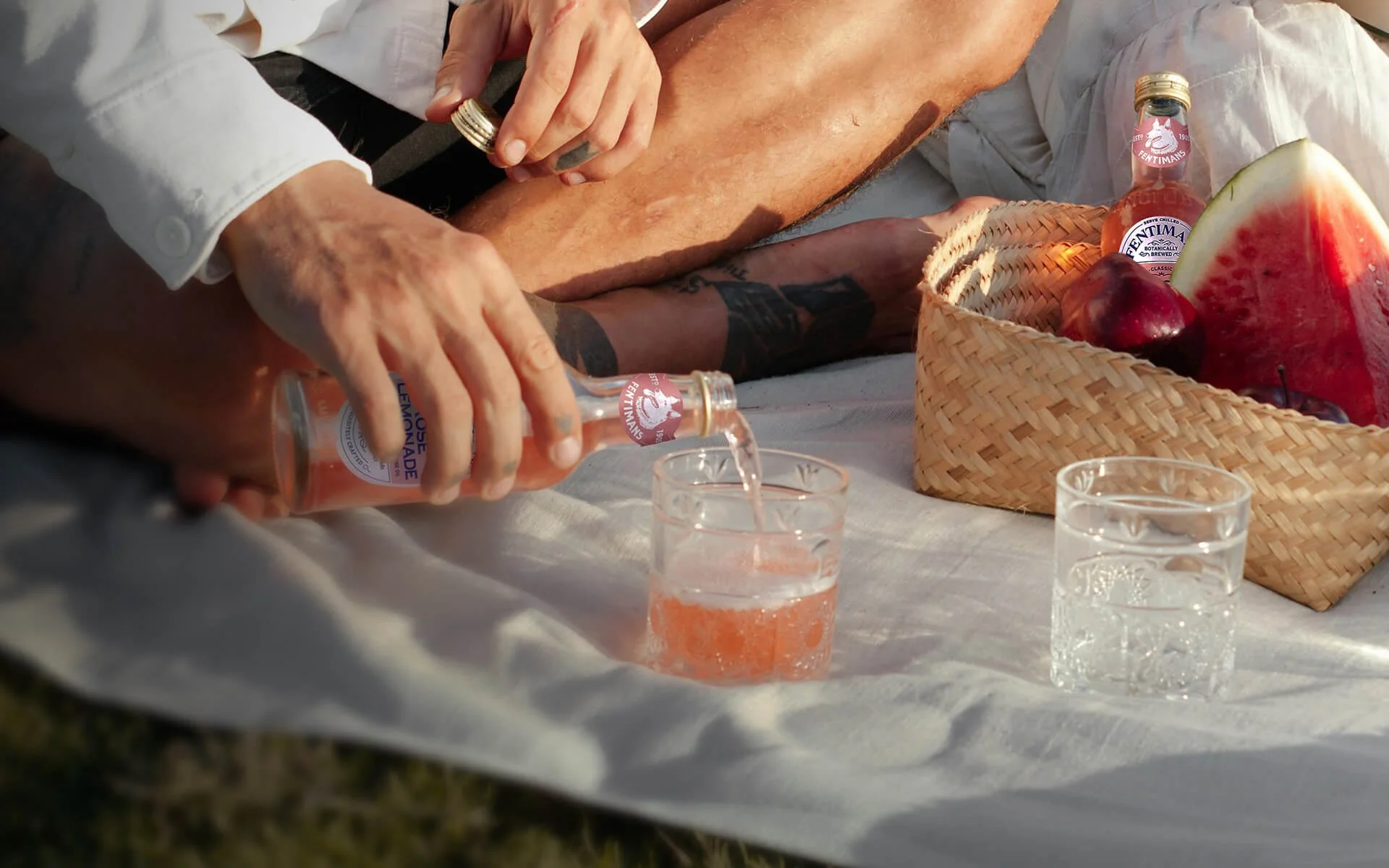 A person in a white shirt is pouring pink water from a glass bottle into a small glass. A basket of watermelon and apples is on the table next to two other glasses filled with water.