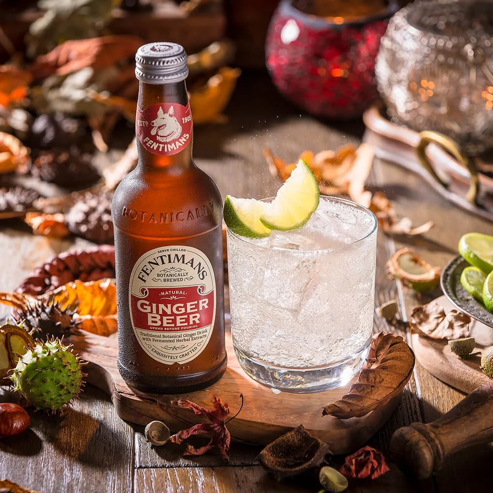 A bottle of Fentimans ginger beer with a glass of ice and lime wedges on a wooden tray surrounded by autumn leaves and decorative items.