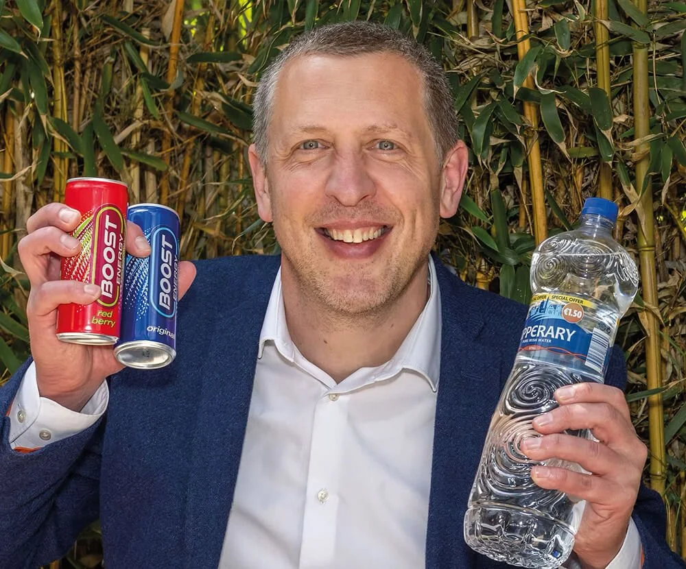 A man in a white shirt and blue blazer holding two cans of Boost energy drink in one hand and a bottle of bottled water in the other, standing in front of a background of green bamboo.
