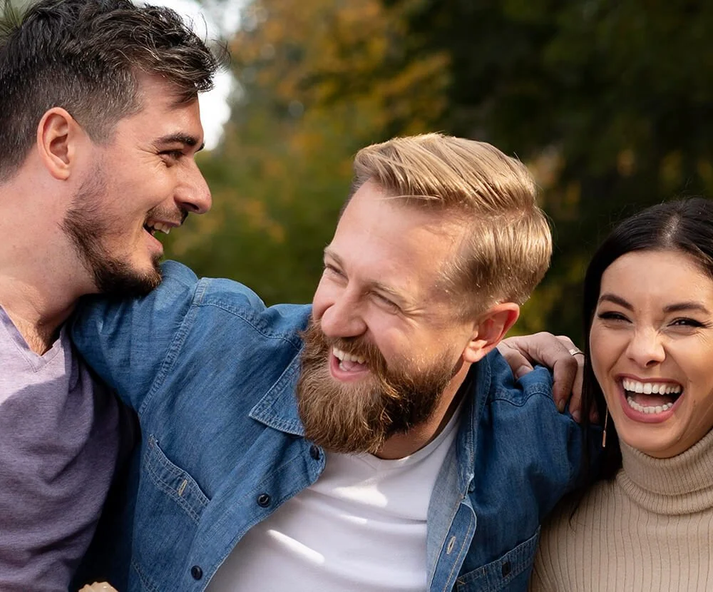 Four smiling friends laughing and embracing outdoors in a park with trees in the background.