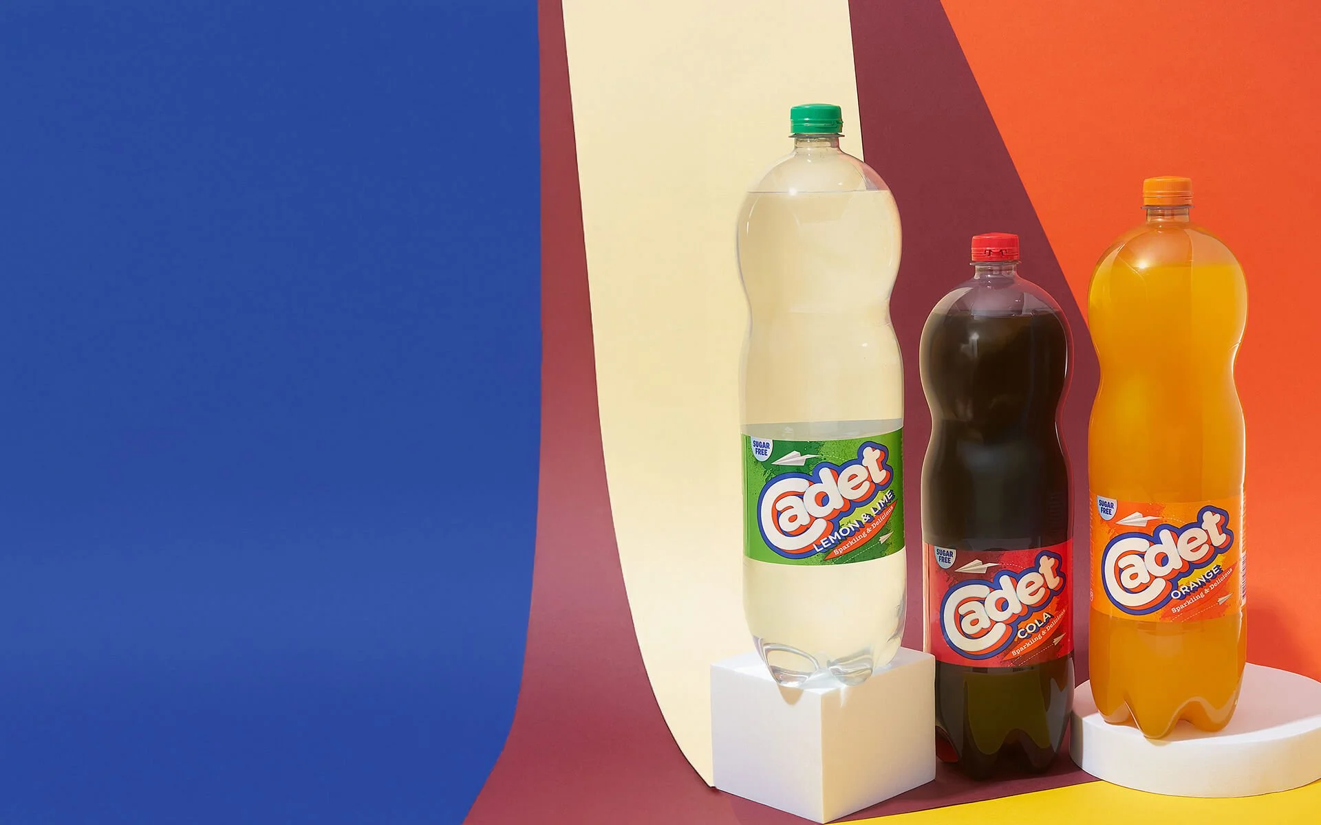 Three bottles of flavored soda soda sit on display against colorful abstract background. The bottles are labeled Adet lemonade lime, cola, and orange, with the lemonade lime on the left, cola in the middle, and orange on the right.