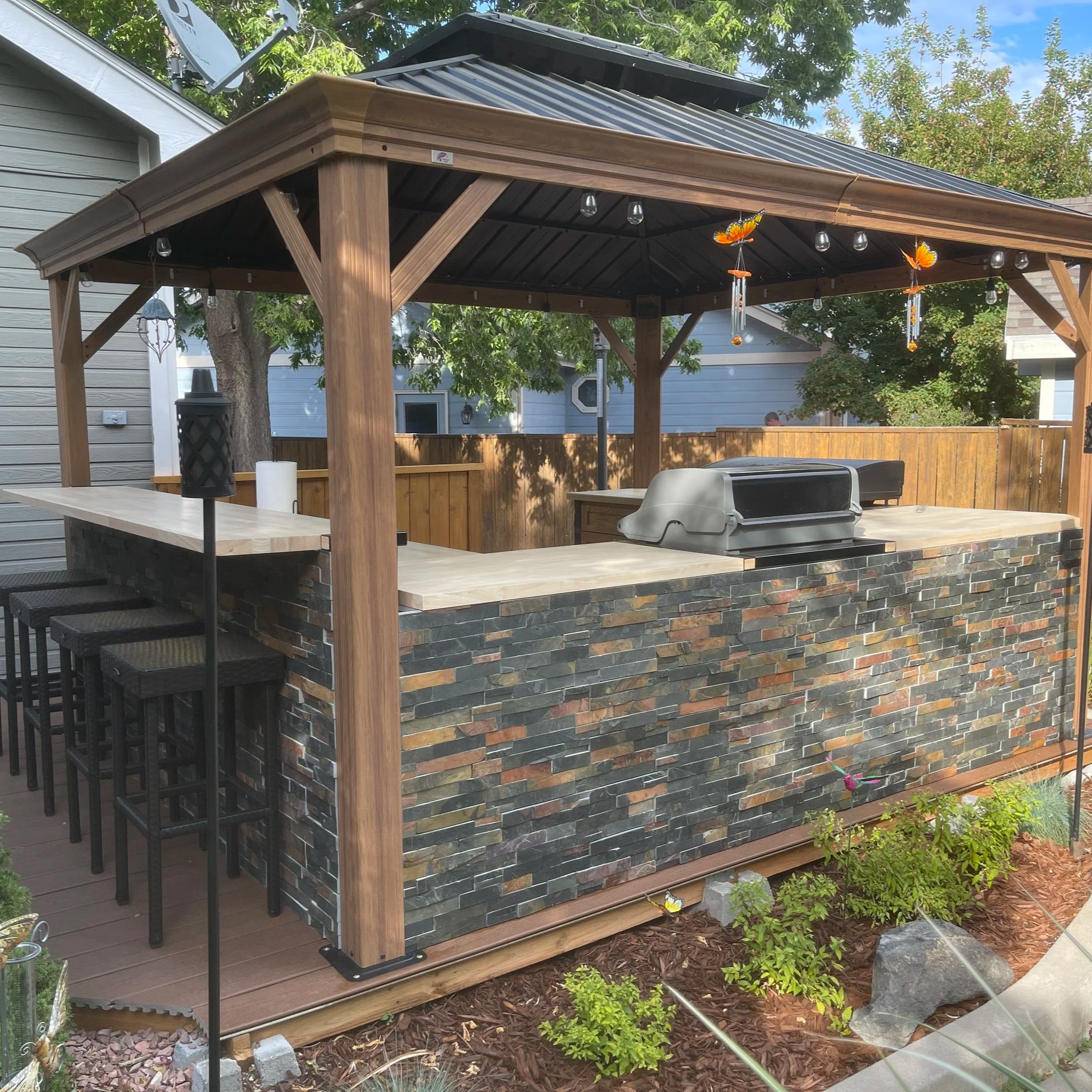 Outdoor kitchen with a stone front, bar stools on a wooden deck, a grill, a lantern, and a shaded wooden pergola with decorative butterflies hanging from the ceiling.