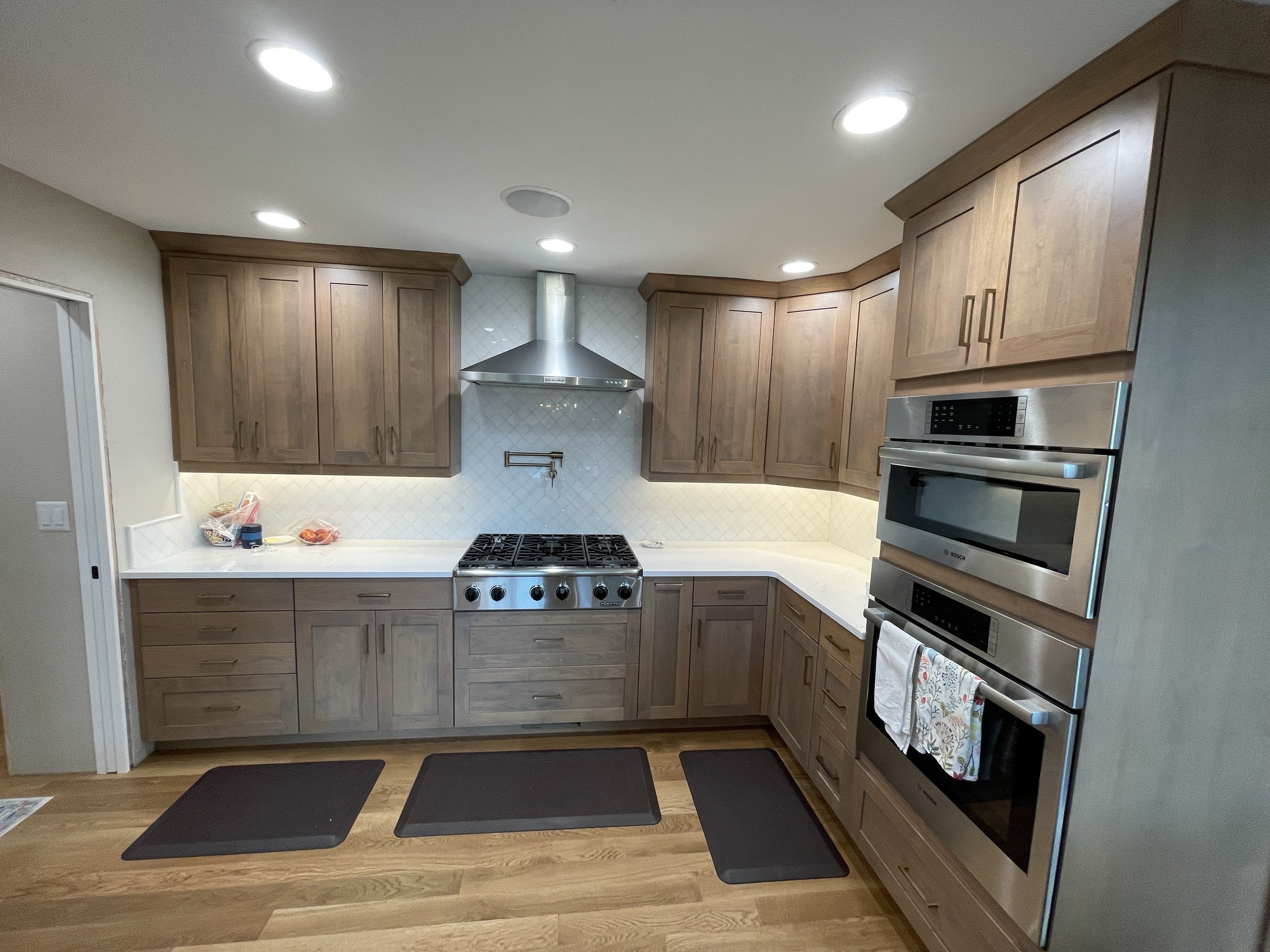 Kitchen with wooden cabinets, white countertops, stainless steel appliances including double oven and gas stove, hardwood floors, three black mats on the floor, and some items on the countertop.