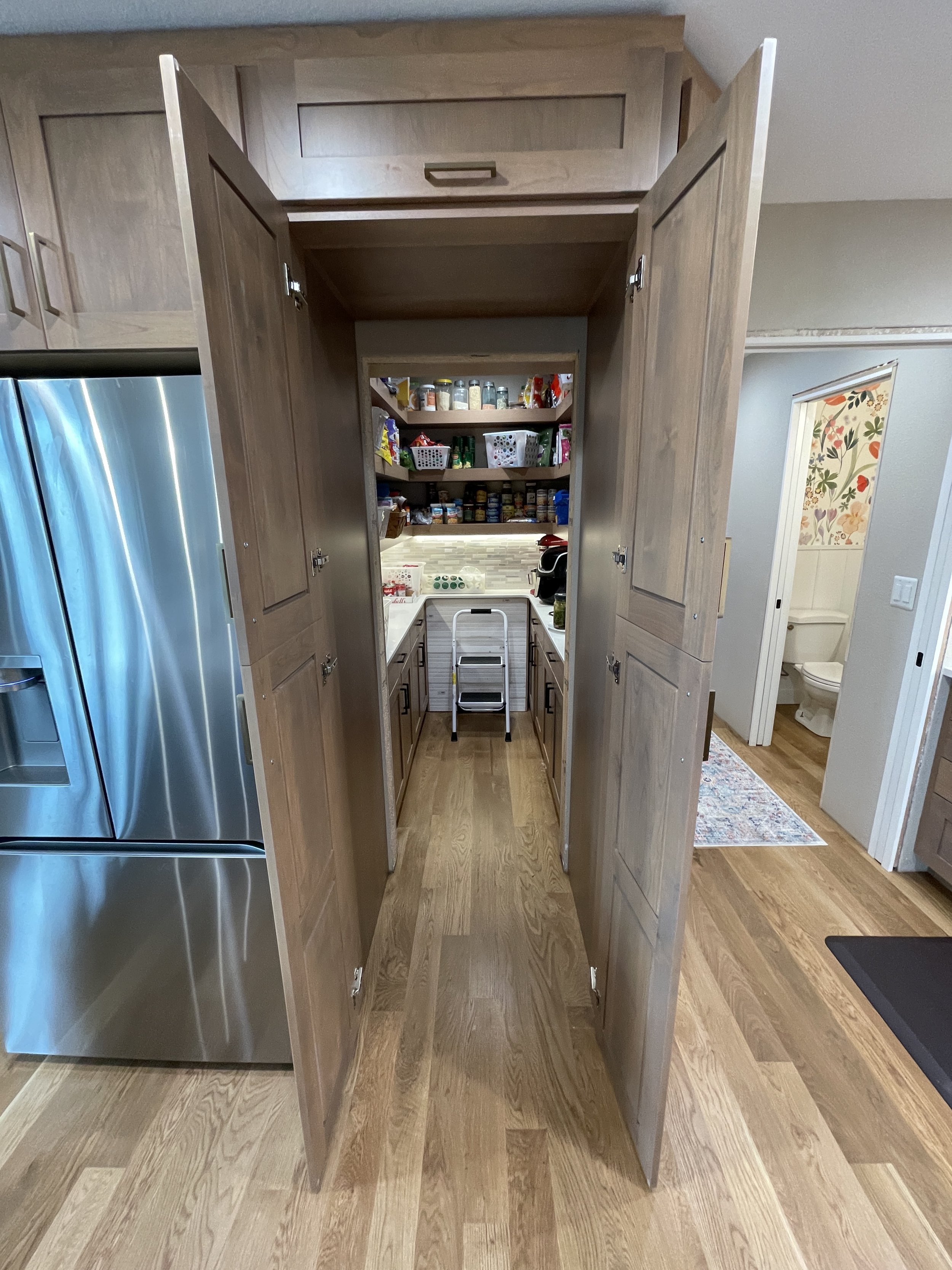 View through open cabinet doors revealing a pantry with shelves stocked with jars, cans, and snack items, and a small step ladder inside.