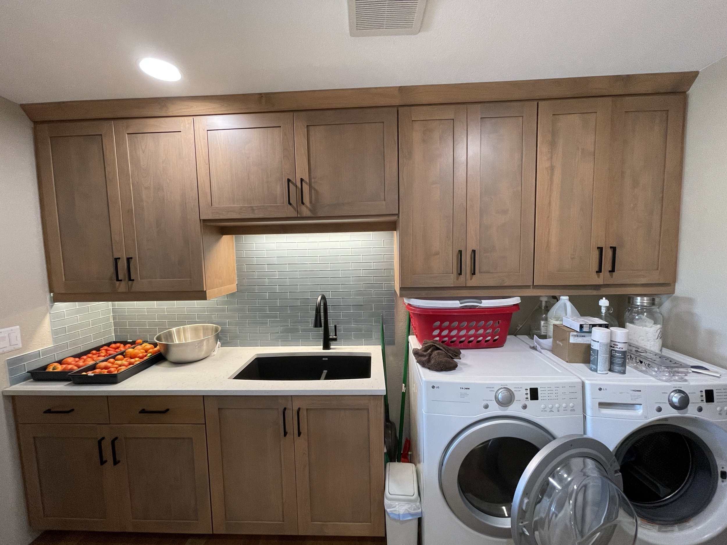 A laundry room with wooden cabinets, a black sink, a countertop with tomatoes and a silver bowl, a red laundry basket, cleaning supplies, and a washing machine and dryer.