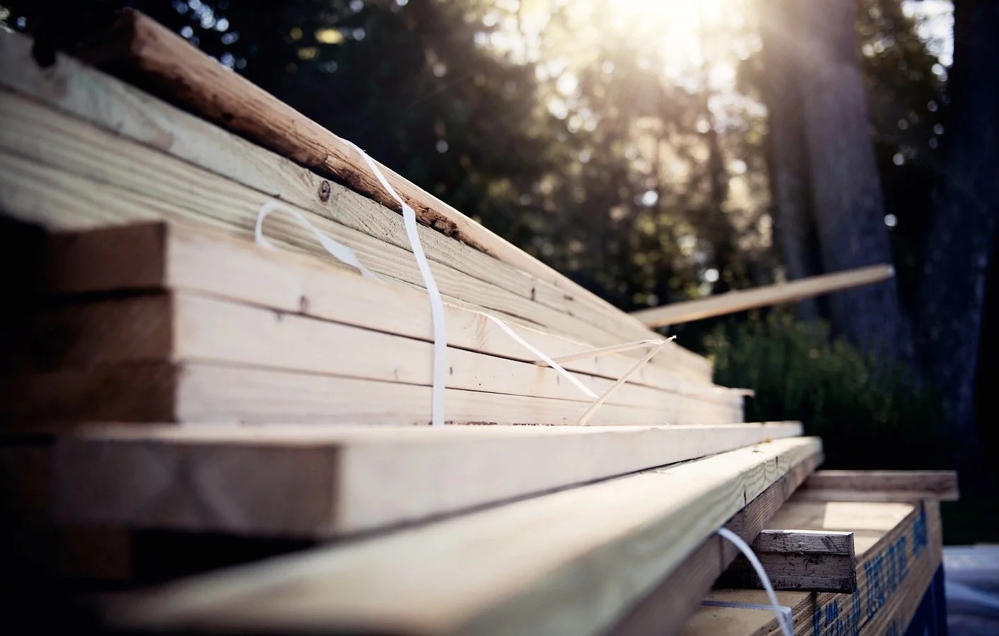 Close-up of stacked wooden boards tied with white plastic straps outdoors with sunlight filtering through trees in background.