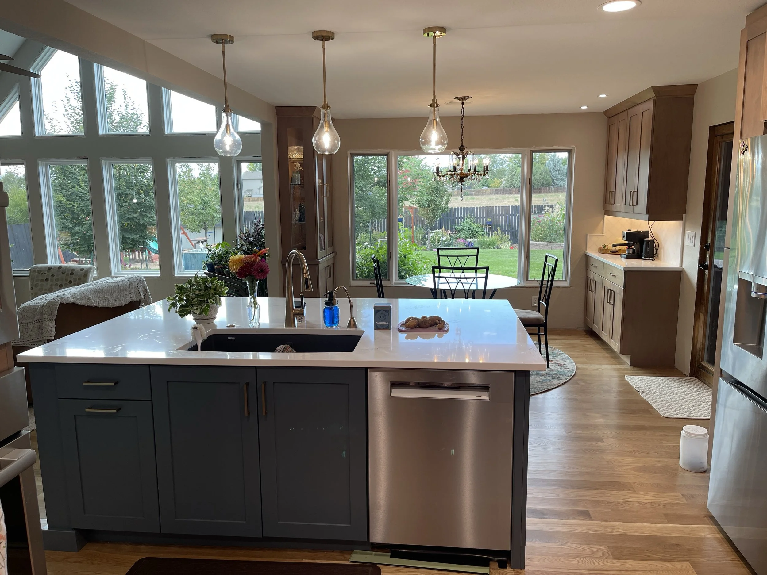Bright kitchen with large windows, island with sink, flowers, and stools, and a dining area with a round table and chairs. Wooden cabinets, stainless steel appliances, and hardwood floors.