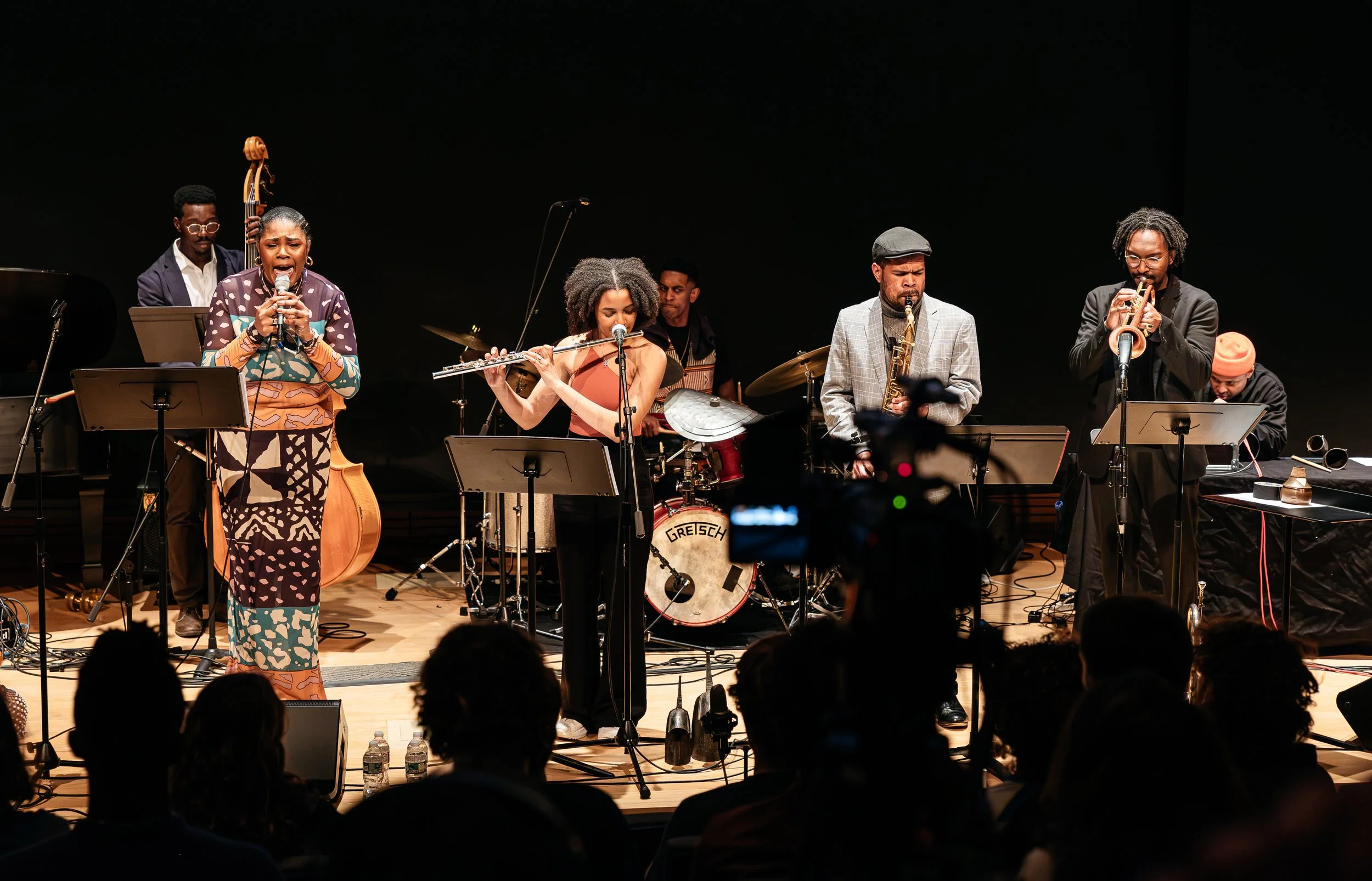 Jazz band performing on stage, with a female singer holding a microphone, a flutist, a drummer, and other musicians playing instruments, audience watching.