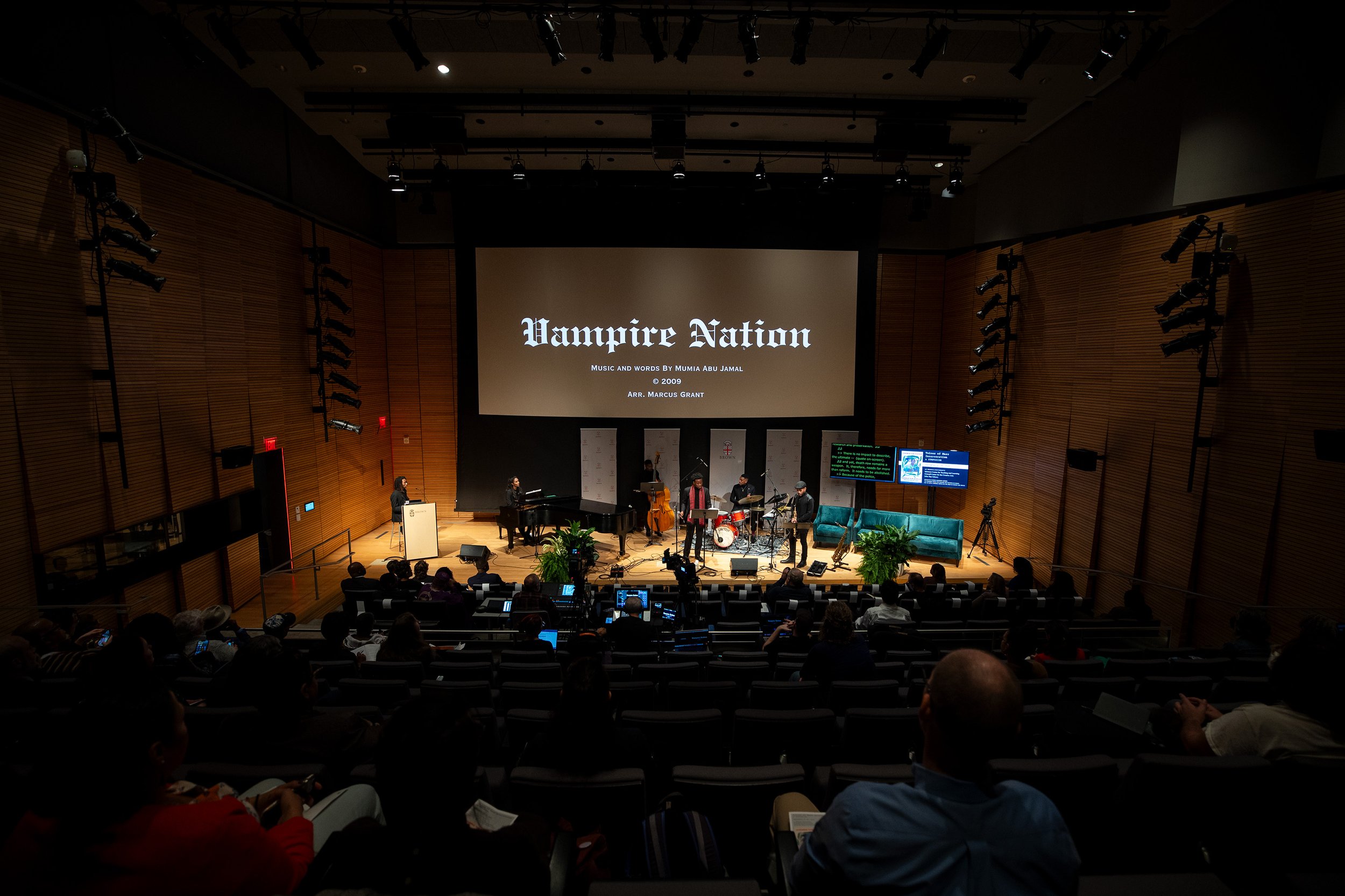 Concert hall audience watching a musical performance on stage with musicians, piano, drums, and speakers, and a large screen displaying the title 'Vampire Nation'.