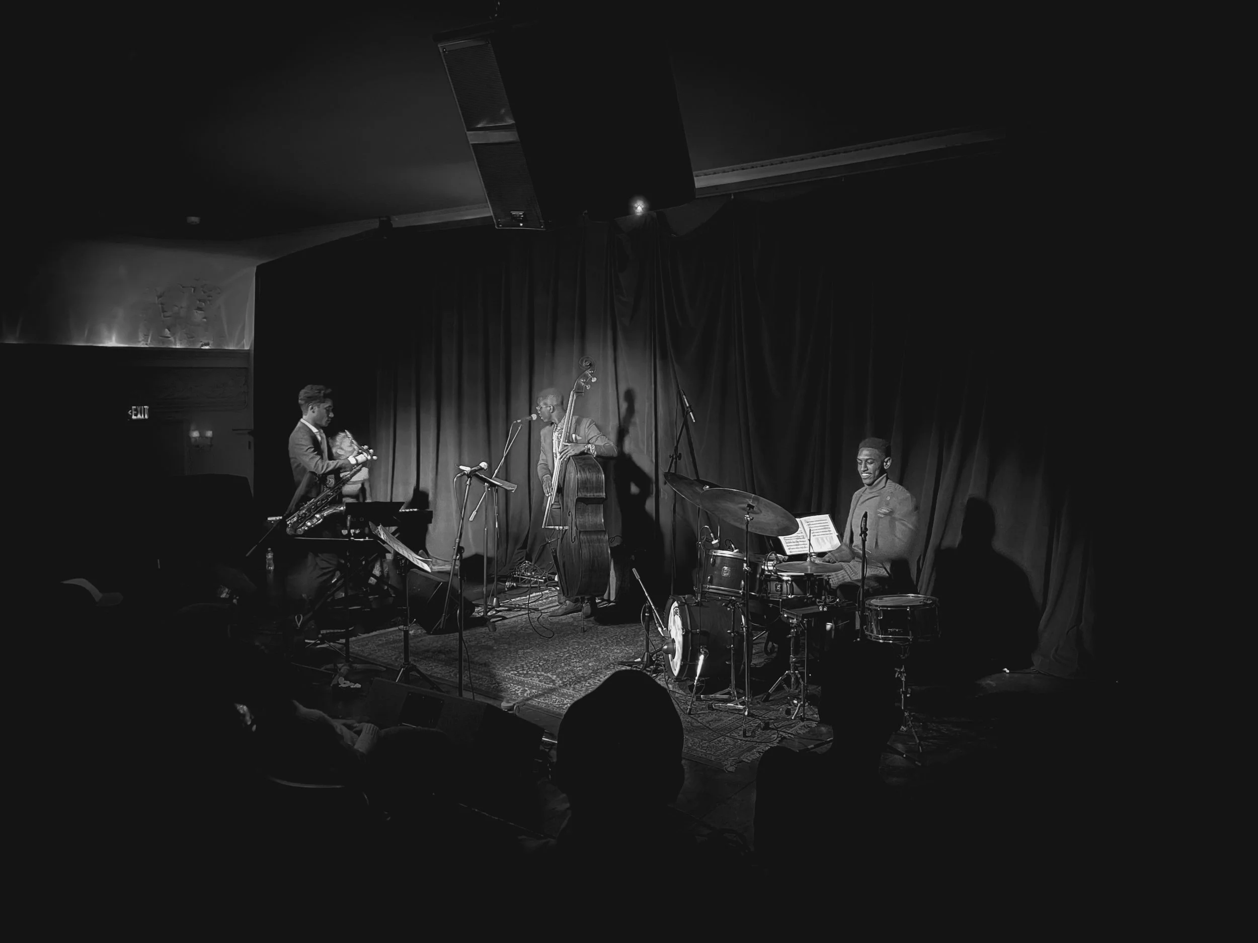 Black and white photo of a jazz band performing on stage with a drummer, double bassist, pianist, and saxophonist, in front of a dark curtain