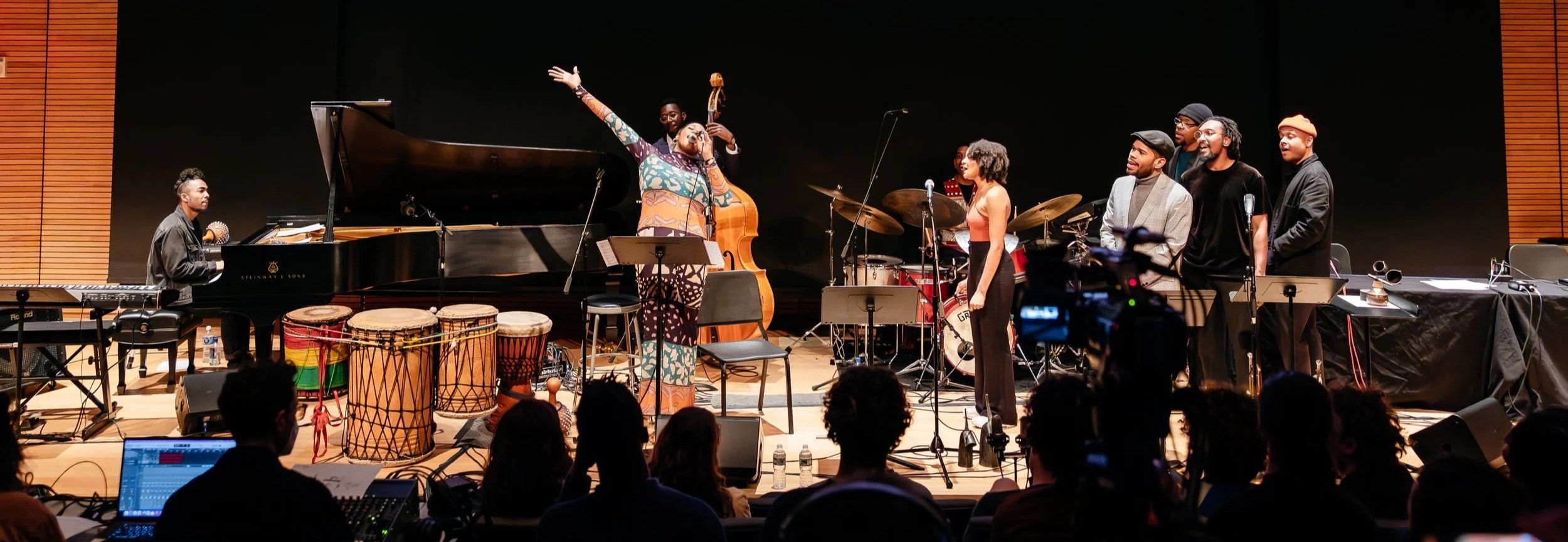 A jazz band performing on stage with several musicians, including a vocalist, pianist, drummer, and others, in a concert hall with audience members visible in the foreground.