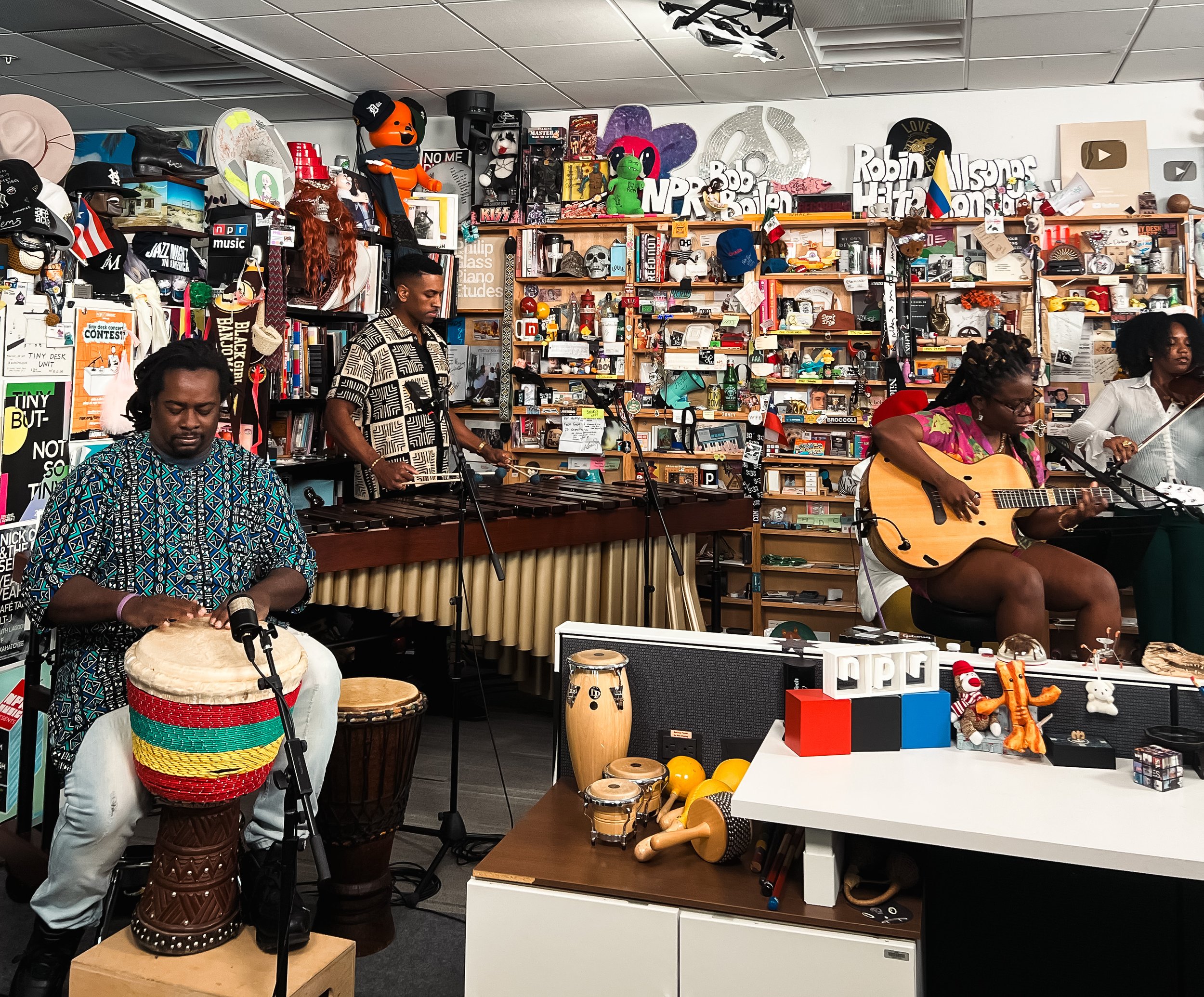 A group of musicians performing in a cozy room filled with eclectic decorations and collectibles, including plush toys, books, art pieces, and musical instruments.
