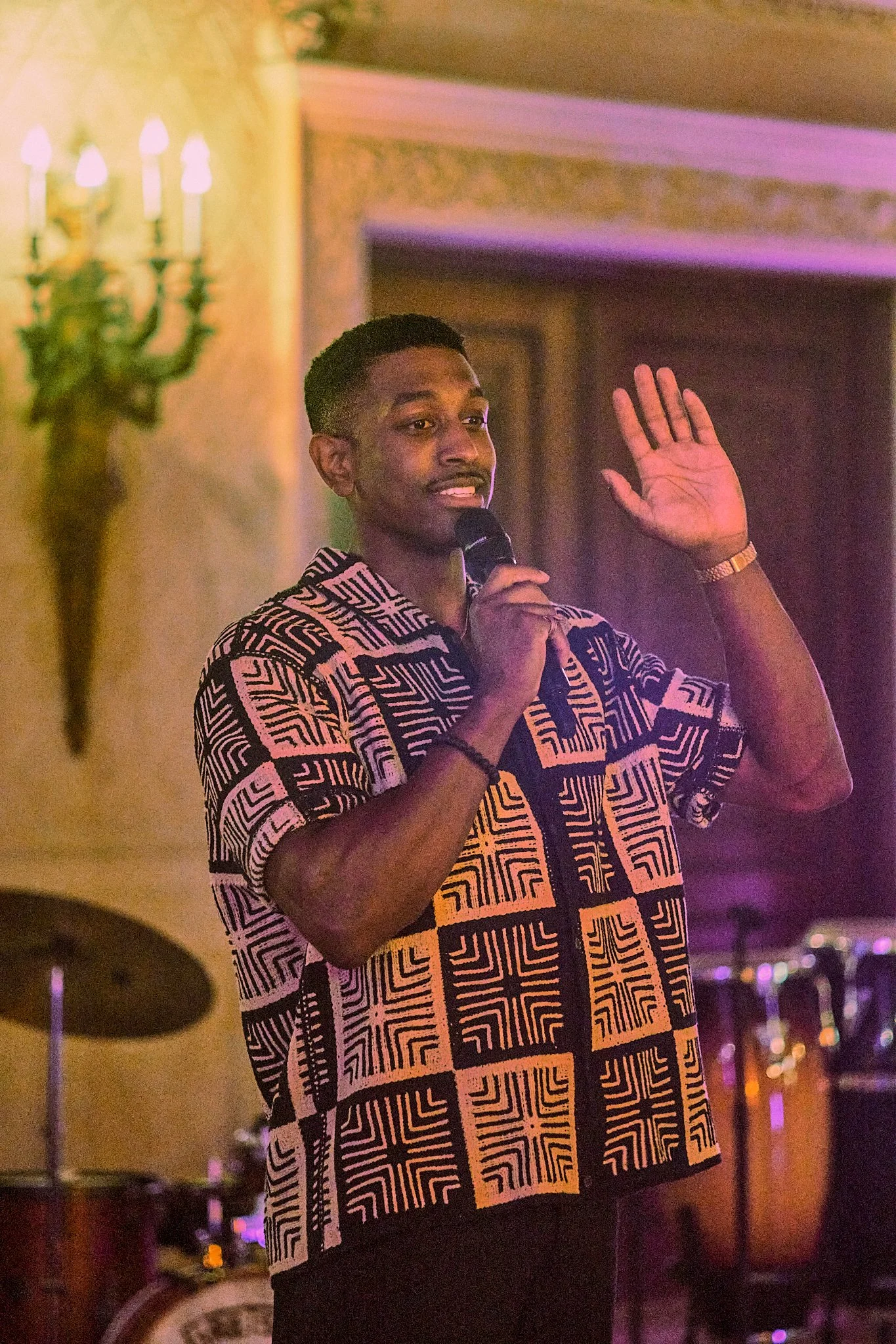 A young man with short black hair and a patterned black and beige shirt is speaking into a microphone and raising his right hand, while standing in a dimly lit room decorated with a wall sconce and purple and pink lighting.