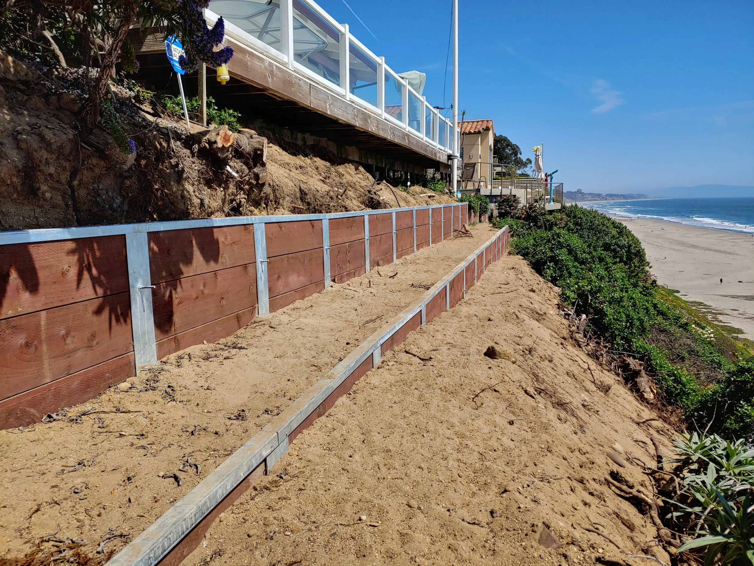 Construction work on a stop at a beachside house, with a wooden and metal retaining wall along the sand, overlooking the ocean.